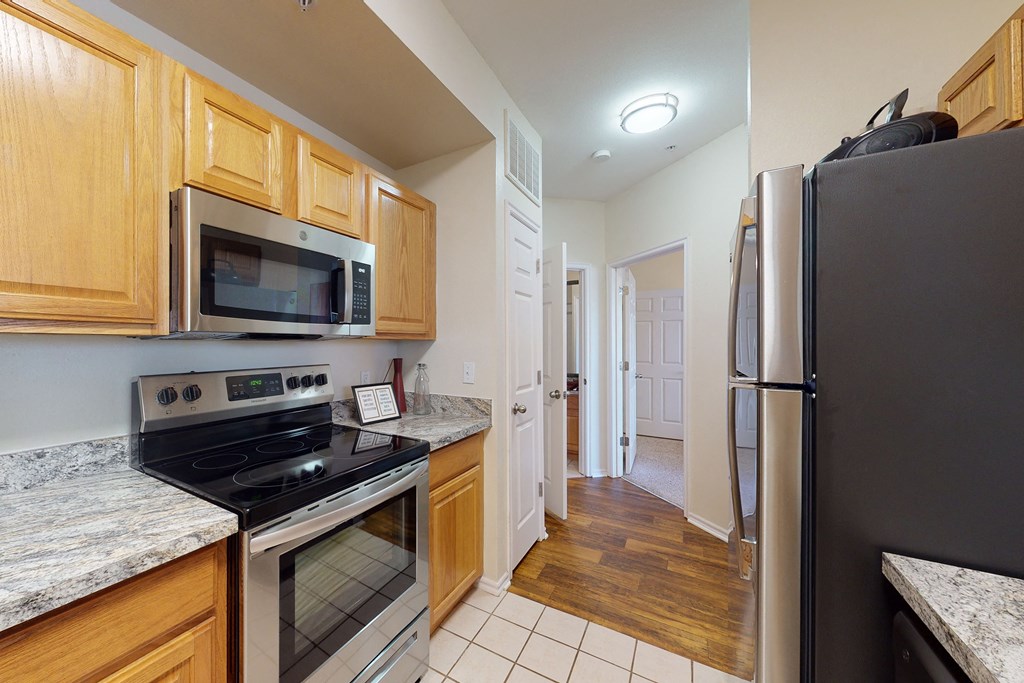Modern kitchen here at Quail Landing Apartment Homes with light wood cabinets, stainless steel appliances, and granite countertops. The floor transitions from tile to wood leading to an open door.