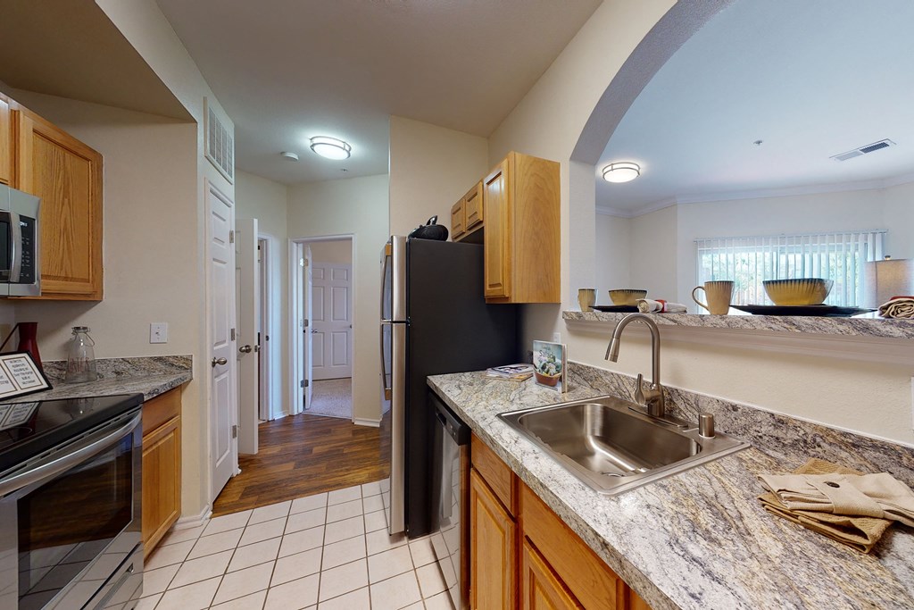 Bright kitchen here at Quail Landing Apartment Homes with wooden cabinets and granite countertops. Stainless steel appliances and a double sink are present. A hallway and living room are visible.