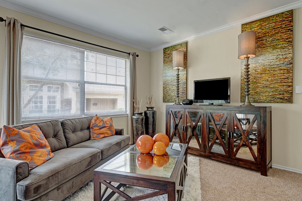 A cozy living room here at Quail Landing Apartment Homes with a gray sofa adorned with vibrant orange pillows, a glass coffee table with orange decor, and a TV on a wooden console.