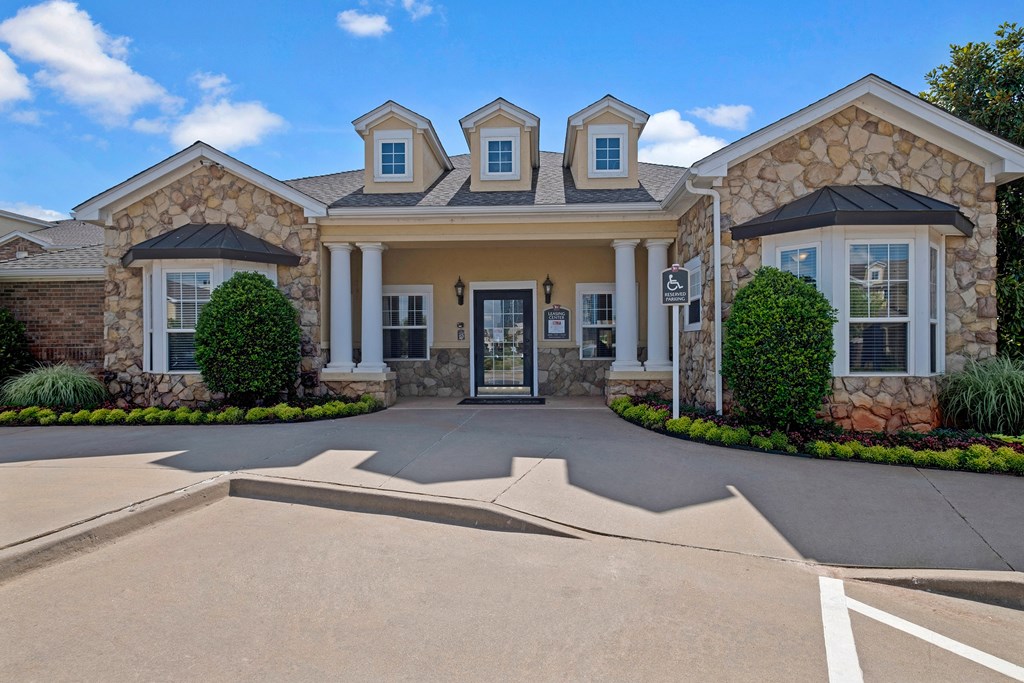 Single-story leasing office here at Quail Landing Apartment Homes with stone facade and three dormer windows. Entrance has two white columns. Surrounded by green shrubs under a blue sky.