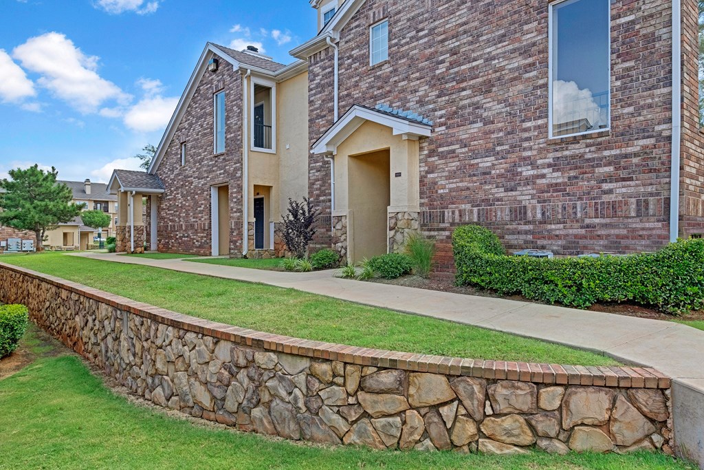 A brick and stone Quail Landing Apartment Homes building with manicured lawns and a stone retaining wall under a bright blue sky. The scene conveys a serene suburban vibe.