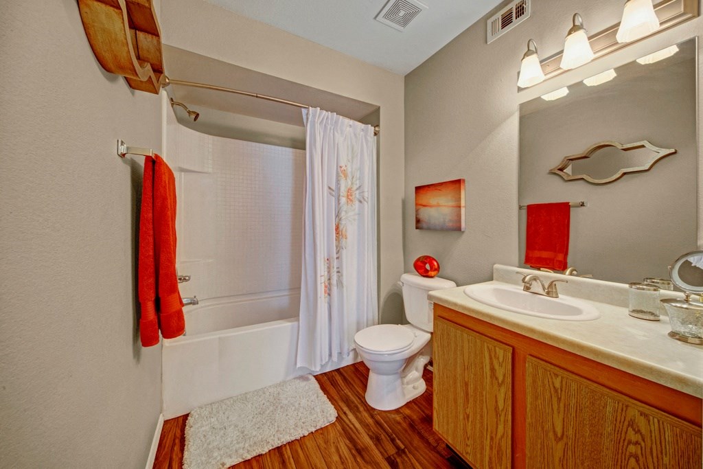 A warm-toned bathroom here at Summit Point Apartment Homes features a shower with a white curtain, a wooden vanity with sink, orange towels, artwork, and soft lighting, creating a cozy atmosphere.