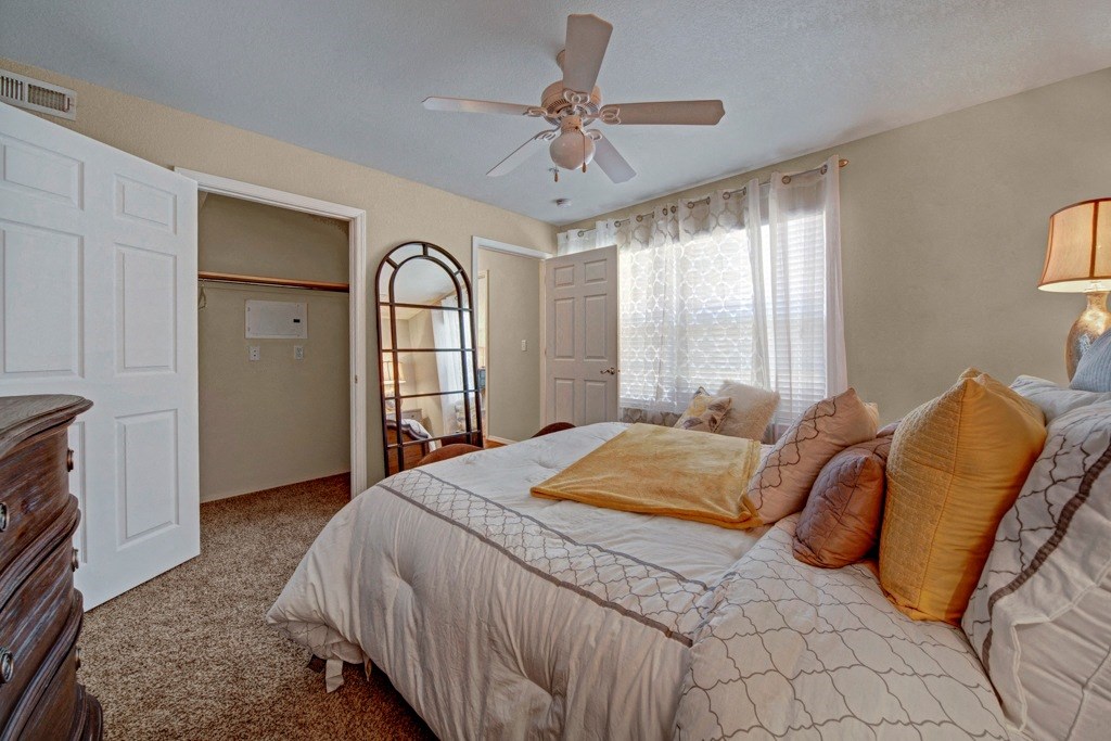 Cozy bedroom here at Summit Point Apartment Homes with neutral tones, featuring a bed with gold and brown pillows, a ceiling fan, an open closet, a large window with light curtains, and a wooden dresser.