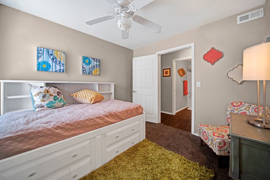 Cozy bedroom here at Summit Point Apartment Homes with a twin bed featuring white built-in drawers, colorful pillows, and abstract wall art. Warm lighting and patterned chair add charm.