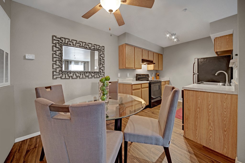 Modern kitchen and dining area here at Summit Point Apartment Homes with gray walls, wood cabinets, and a ceiling fan. Features a glass table, beige chairs, and decorative mirror.