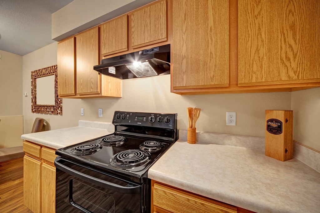 Kitchen scene here at Summit Point Apartment Homes with oak cabinets, a black stove, and a beige countertop. Wooden utensils in a holder, framed mirror, and inviting, cozy ambiance.