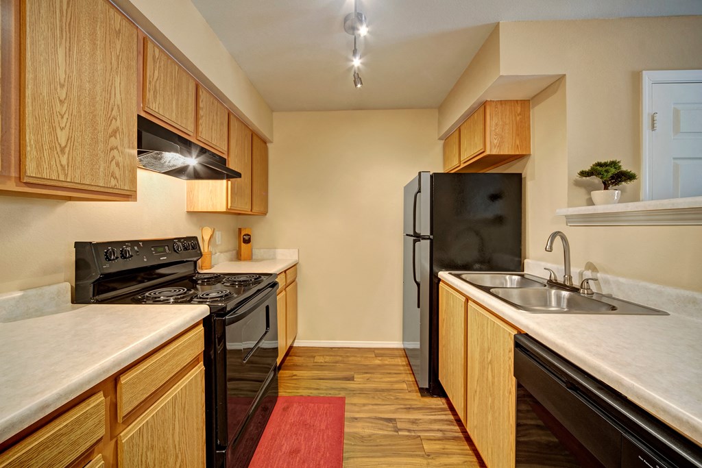 Compact kitchen here at Summit Point Apartment Homes with light wood cabinets, black appliances, and laminate flooring. Features a double sink, track lighting, and a small potted plant. Cozy ambiance.
