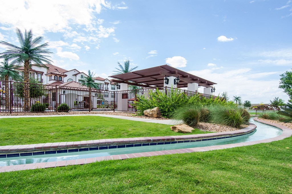 Beautiful outdoor scene here at Andalucia Villas Apartment Homes with a lazy river curving through a lush lawn, bordered by palm trees and rocks. A pergola and bright sky add tranquility.