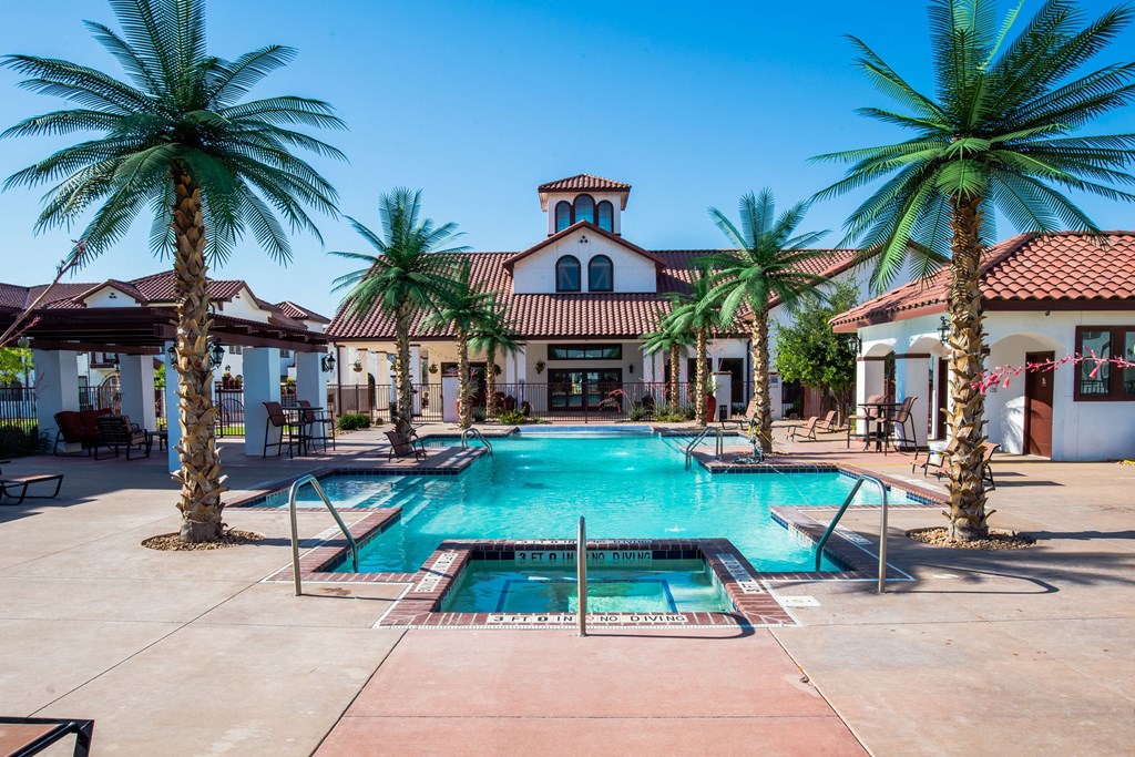 A tranquil resort pool area here at Andalucia Villas Apartment Homes with palm trees under a clear blue sky. A hot tub sits in the foreground, with a Spanish-style building in the background.