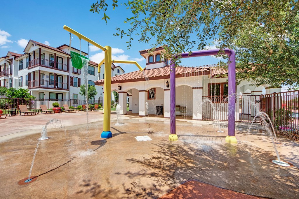 Outdoor splash pad here at Andalucia Villas Apartment Homes with colorful yellow and purple structures spraying water, set against a backdrop of modern apartments and blue sky, evoking playful fun.