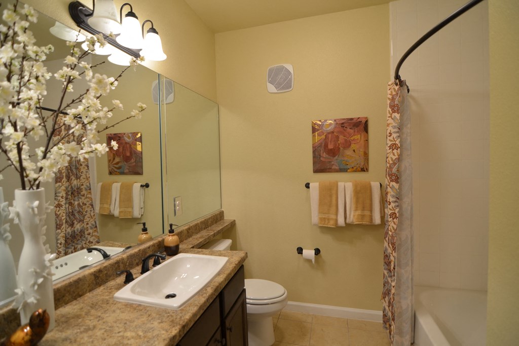 Cozy bathroom here at Andalucia Villas Apartment Homes with warm tones features a granite countertop, white sink, floral arrangement, art, and floral shower curtain. Bright, inviting ambiance.