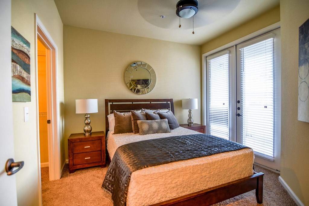 Cozy bedroom here at Andalucia Villas Apartment Homes with a wooden bed, gray and white bedding, and two nightstands with lamps. A round mirror hangs above the bed. Bright natural light from patio doors. Neutral tones create a calm, inviting atmosphere.