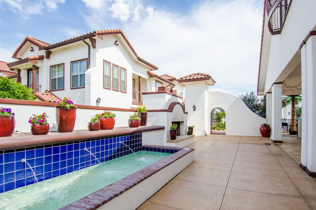 Spanish-style courtyard here at Andalucia Villas Apartment Homes with a calming blue-tiled fountain, terracotta potted flowers, and white stucco buildings with red tiled roofs under a bright sky.