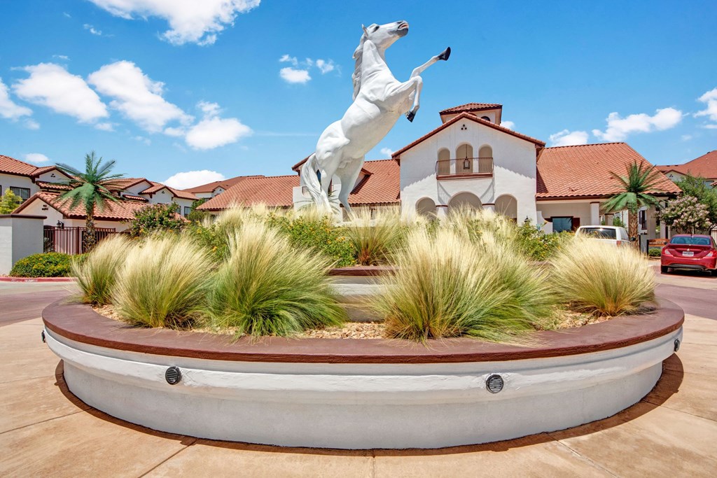 A majestic white horse statue rears on a large circular platform adorned with tall grasses here at Andalucia Villas Apartment Homes. In the background, a building with terracotta roofs under a clear blue sky.