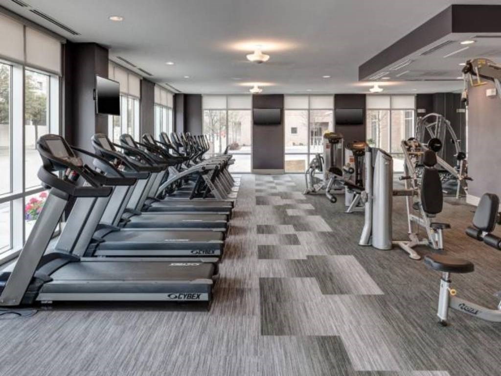 Modern gym here at Arpeggio Apartment Homes with rows of treadmills on the left and strength training machines on the right. Large windows provide natural light, creating an inviting atmosphere.