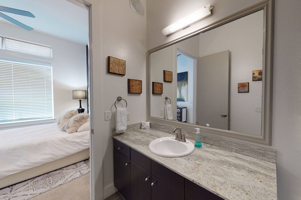 Modern bathroom here at Arpeggio Apartment Homes with a large mirror, granite countertop, and sink. A glimpse into a cozy bedroom with a bed, pillows, and a lit lamp. Decor is minimalist.