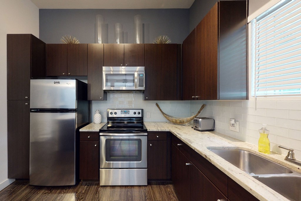 Modern kitchen here at Arpeggio Apartment Homes with dark wood cabinets, stainless steel appliances, white tiled backsplash, and granite countertops. Natural light enters through blinds, creating a cozy ambiance.