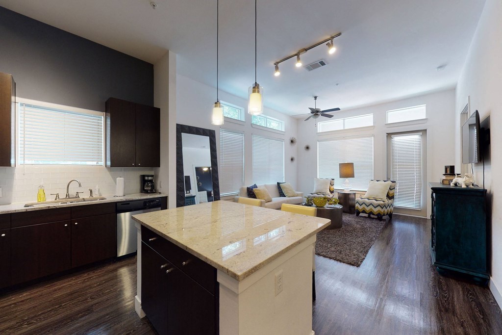Modern open-concept living room and kitchen here at Arpeggio Apartment Homes with dark wood cabinets, marble island, and pendant lights. Cozy seating area, large windows, ceiling fan.