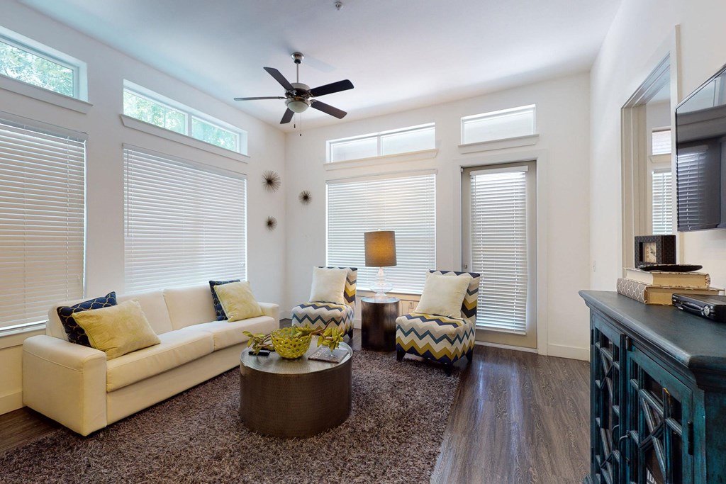 Modern living room here at Arpeggio Apartment Homes with large windows, white sofa, patterned armchairs, and a round coffee table on a plush rug. Ceiling fan and decorative elements create a cozy ambiance.