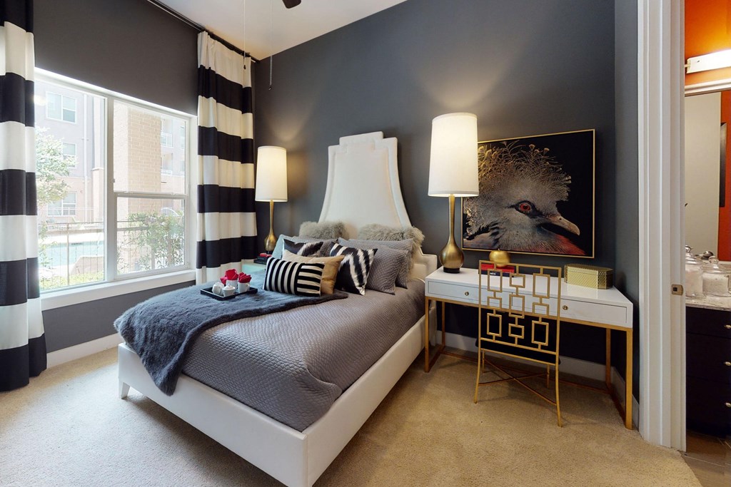 Chic bedroom here at Arpeggio Apartment Homes with gray and white decor, featuring a plush bed with striped pillows. Two gold lamps frame a bird art piece, exuding elegance.
