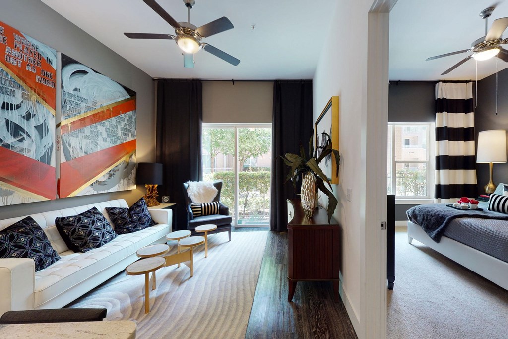 Modern living room here at Arpeggio Apartment Homes with abstract red and gray artwork, white sofa, black and white pillows, and a wooden coffee table. Bedroom visible, elegant decor.