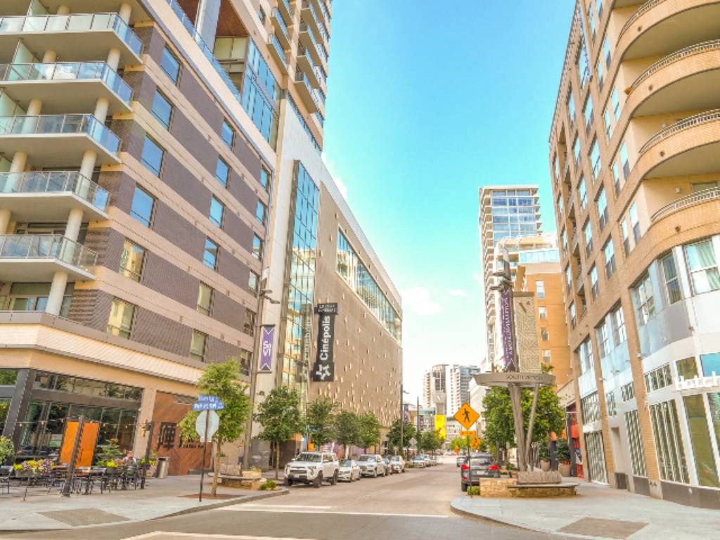 Urban street scene near Arpeggio Apartment Homes with modern high-rise buildings, including outdoor seating and green trees, under a bright blue sky. The atmosphere feels lively and bustling.