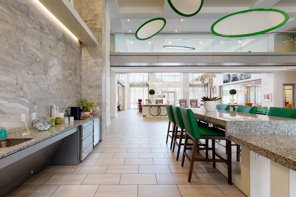 Modern lounge area here at Arpeggio Apartment Homes with a long granite table and green chairs. Circular green ceiling lights and natural light from large windows create a bright, inviting atmosphere.