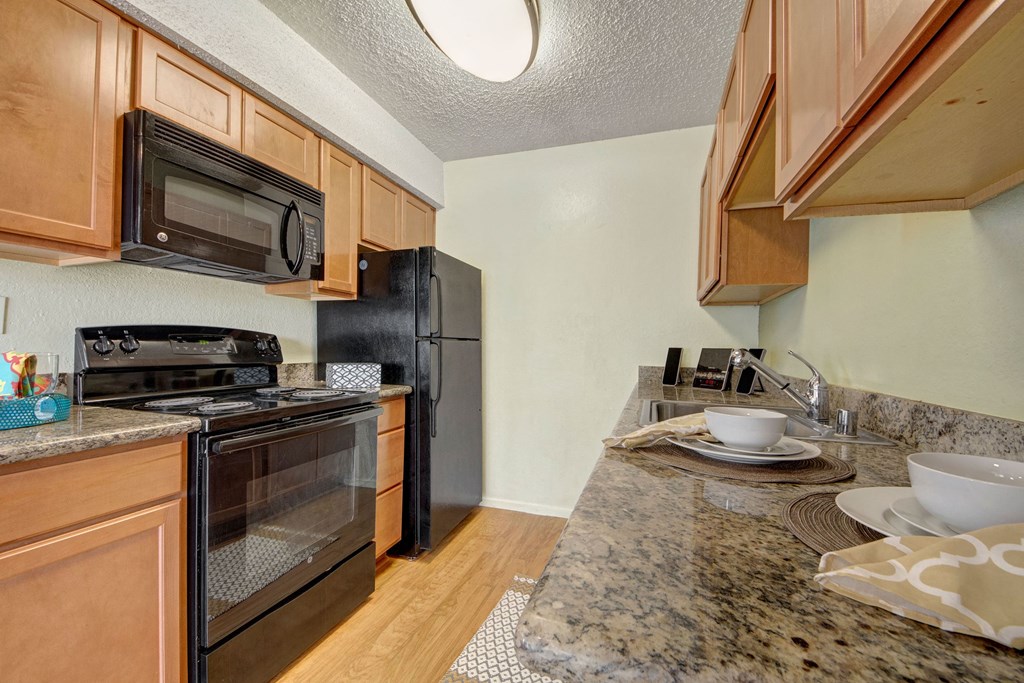 Compact kitchen here at Avalon Springs Apartment Homes with warm-toned wood cabinets, granite countertops, a black stove, microwave, and fridge. Light wood flooring and a cozy atmosphere.