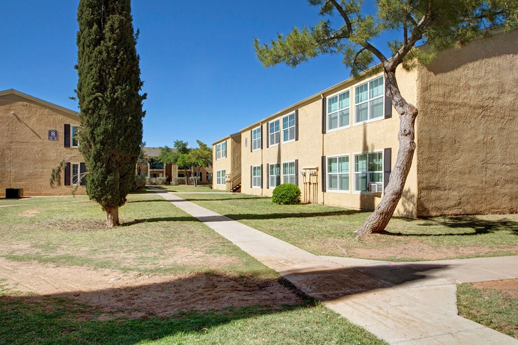 Sunny day at Avalon Springs Apartment Homes with beige two-story buildings, manicured lawns, a tall cypress tree, and a winding concrete path under clear blue skies.