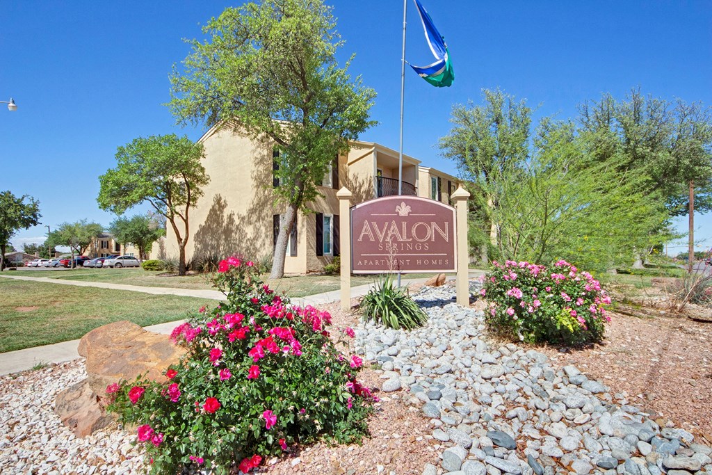 Sign for Avalon Springs Apartment Homes surrounded by vibrant pink flowers and green shrubs. Beige building and clear blue sky in the background convey a serene atmosphere.