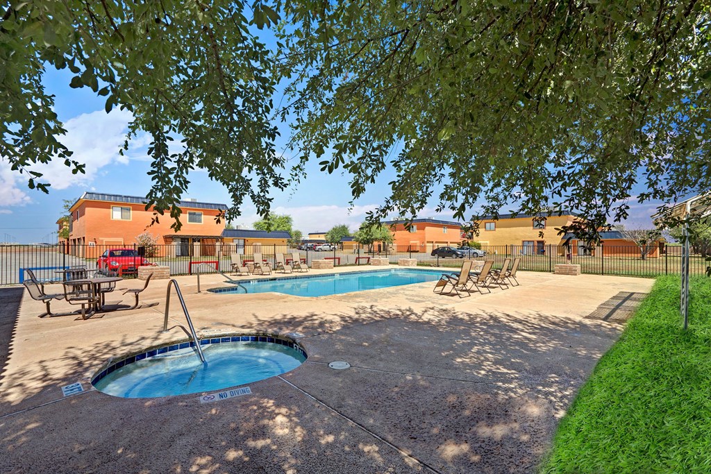 Shaded by trees, a pool area here at Avanti Townhomes with loungers and a hot tub is surrounded by grass and buildings. A tranquil setting under a bright blue sky.