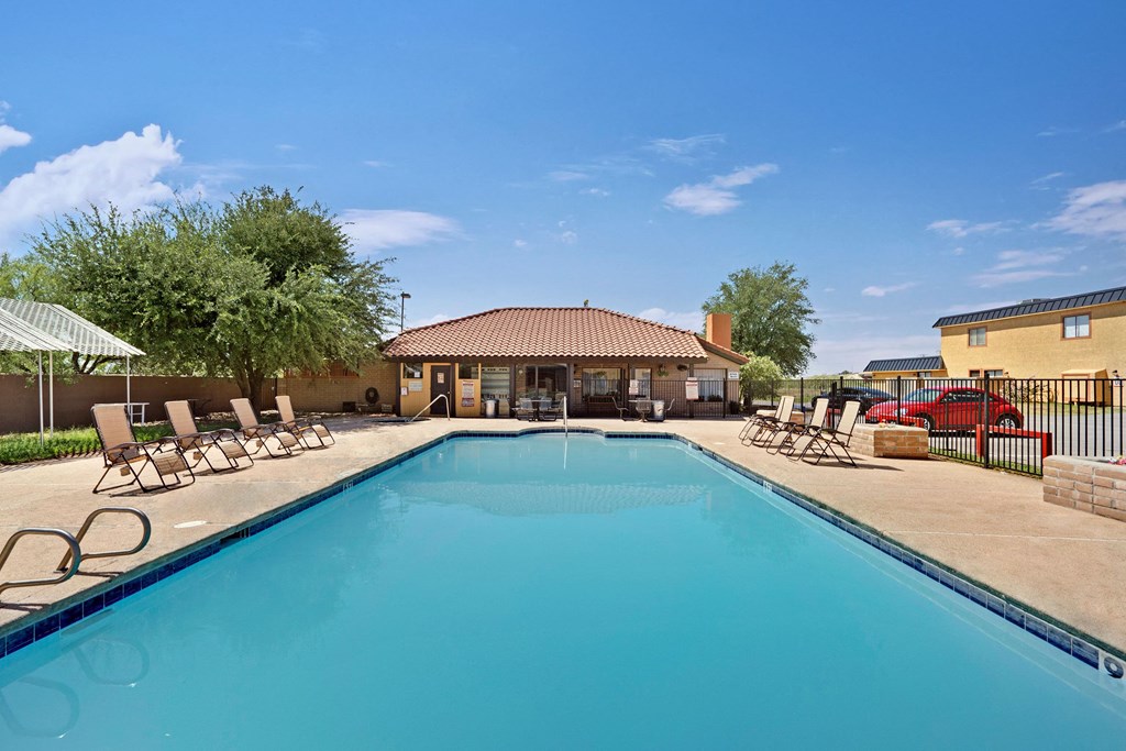 Outdoor pool here at Avanti Townhomes with clear blue water, surrounded by tan lounge chairs under a bright blue sky. A small building with a red-tiled roof is in the background.