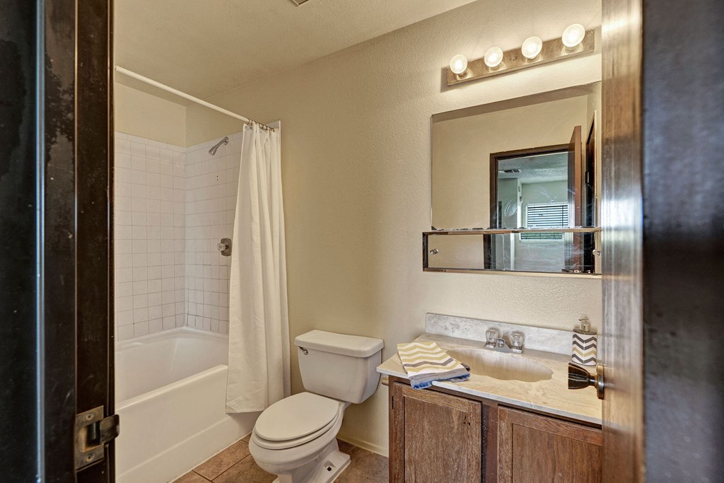 A small bathroom here at Avanti Townhomes with a bathtub and shower, white curtain, toilet, and wooden vanity. A mirror and four-bulb light fixture add brightness.