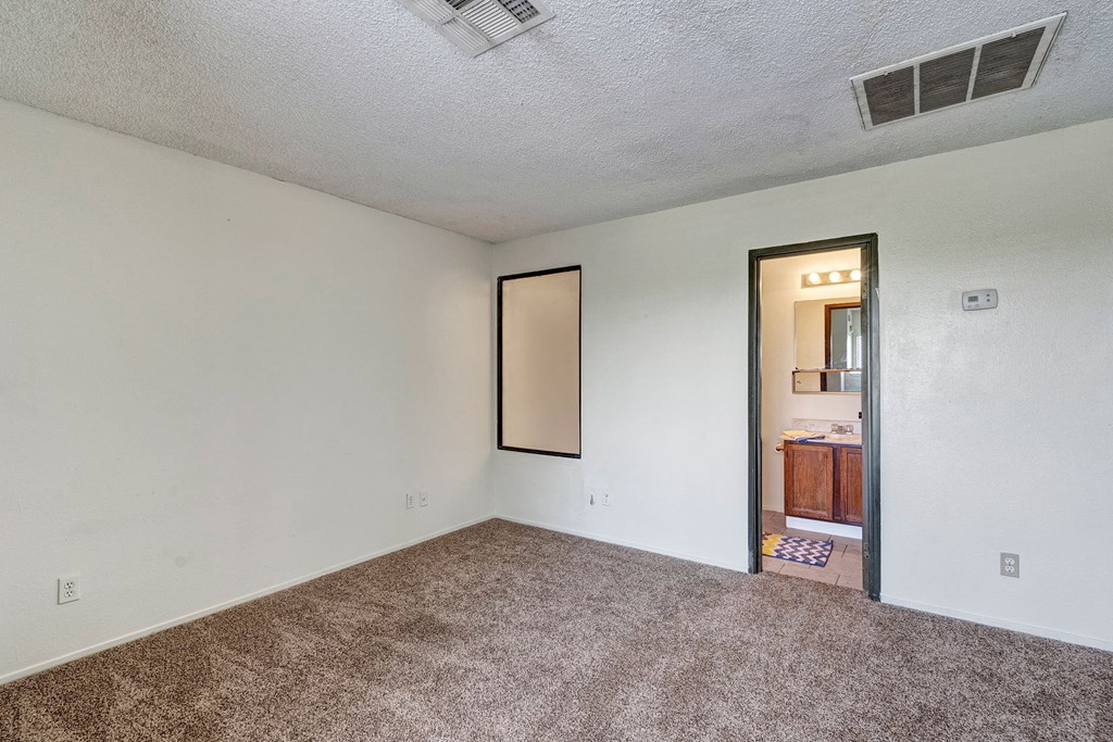Empty bedroom here at Avanti Townhomes with beige carpet and white walls. A door leads to a small bathroom with a lit mirror and wooden cabinets. Calm and neutral ambiance.