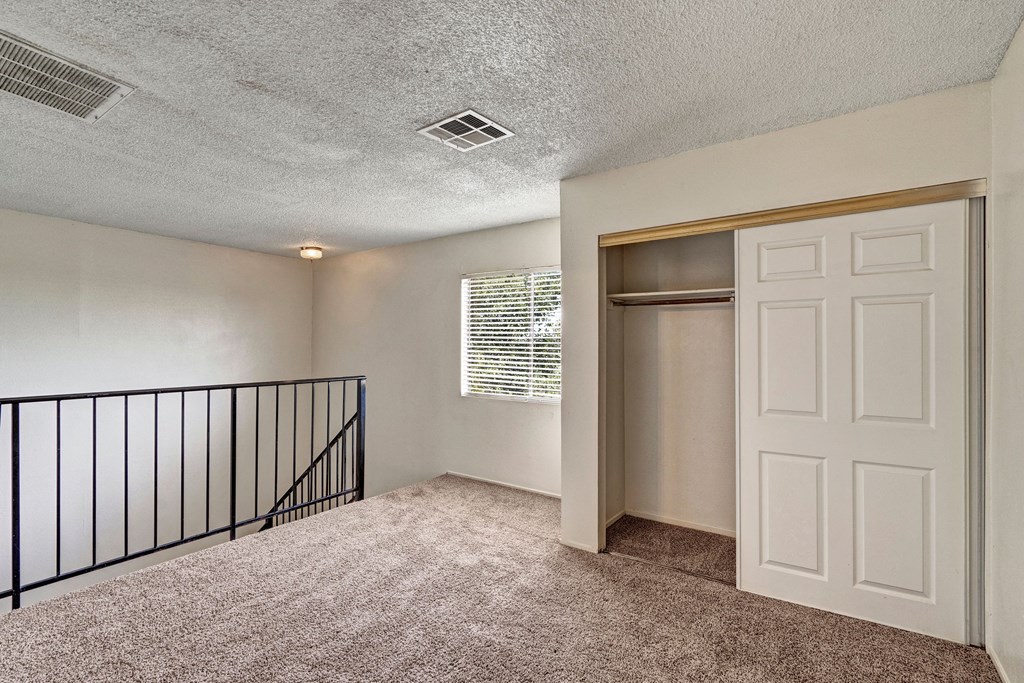 Carpeted room here at Avanti Townhomes with a white closet having open sliding doors. A window with blinds lets in natural light. Black railing and a staircase create a cozy atmosphere.