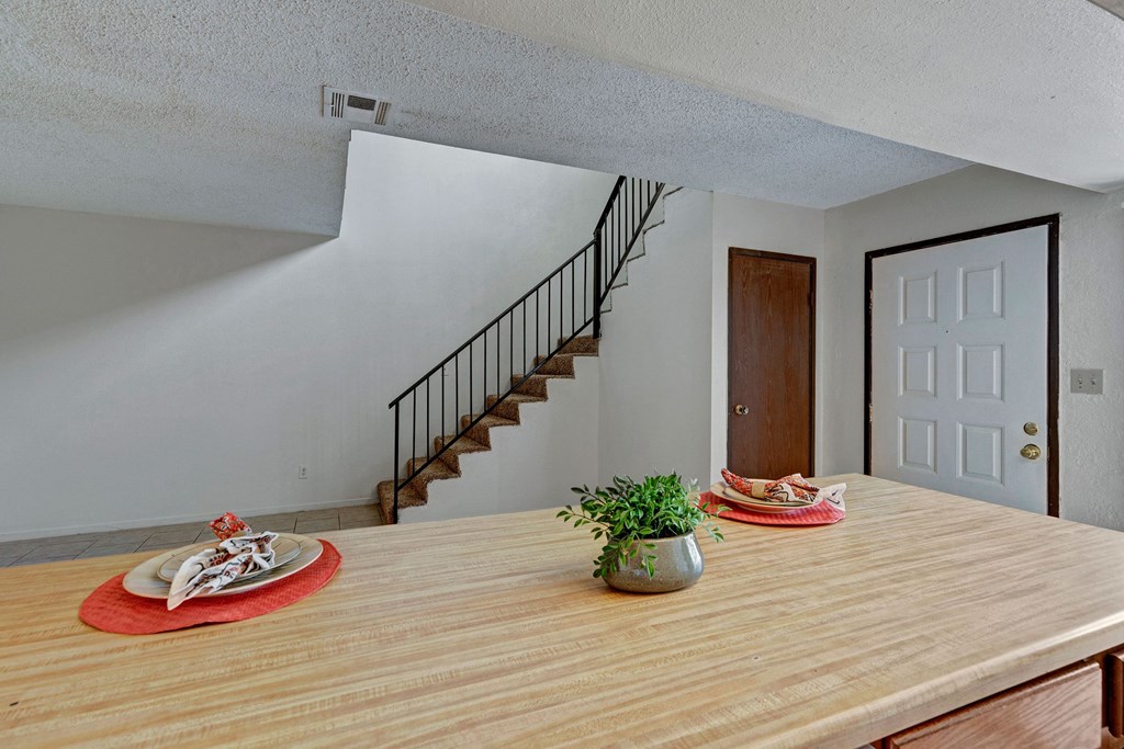 A cozy interior features here at Avanti Townhomes a wooden table with a potted plant and neatly set plates on red placemats. A carpeted staircase with a black railing leads upstairs, creating an inviting, homely ambiance.