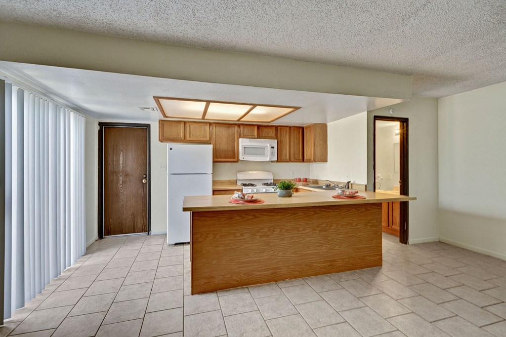 A clean kitchen here at Avanti Townhomes with wooden cabinets and a white fridge features a central island set with red plates and a plant. The room is bright with tiled floors.