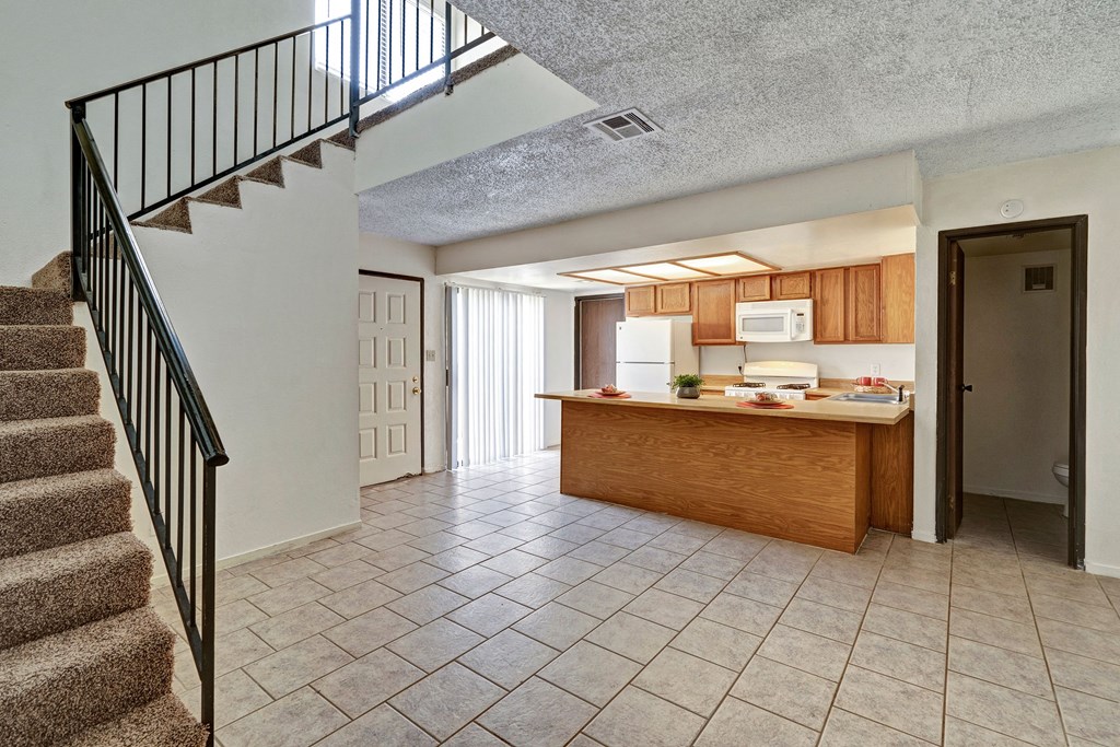 Open kitchen and living area here at Avanti Townhomes with beige tiled floors, wooden cabinetry, and a central island. Carpeted stairs with a black railing lead upstairs. Neutral tones evoke a cozy, welcoming atmosphere.