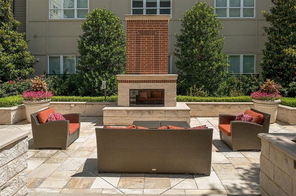 Outdoor patio here at Cityplace Heights Apartment Homes with wicker furniture surrounding a brick fireplace. Greenery and pink flowers in planters add a cozy, inviting atmosphere.