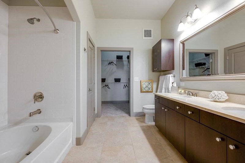 Modern bathroom here at Cityplace Heights Apartment Homes with beige tiles, dark wood vanity, and double sinks. A large mirror and white bathtub enhance the clean, spacious feel.