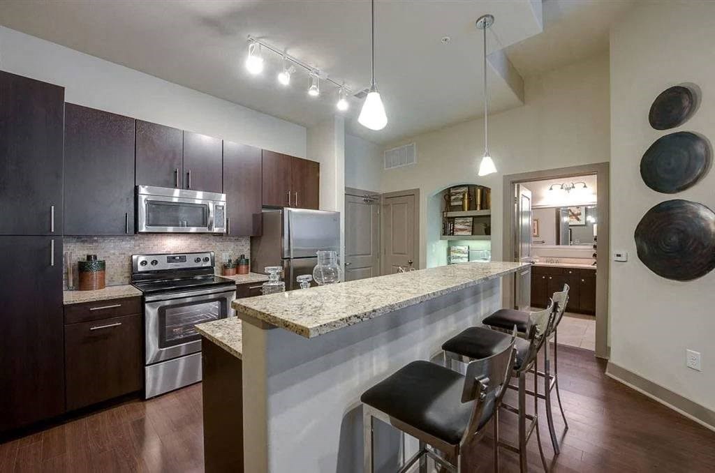 Modern kitchen here at Cityplace Heights Apartment Homes with dark wood cabinets, stainless steel appliances, and a granite countertop bar with four stools. Warm lighting creates a cozy atmosphere.
