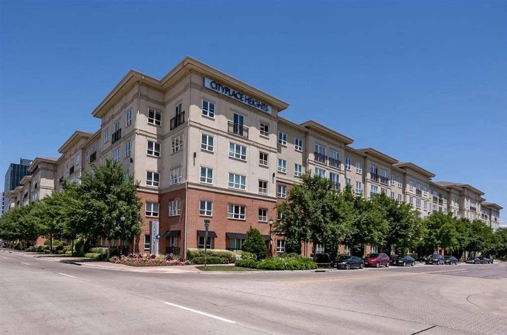 Four-story Cityplace Heights Apartment Homes building with tan and red brick facade, surrounded by lush trees. Bright blue sky adds a peaceful tone to the urban scene.