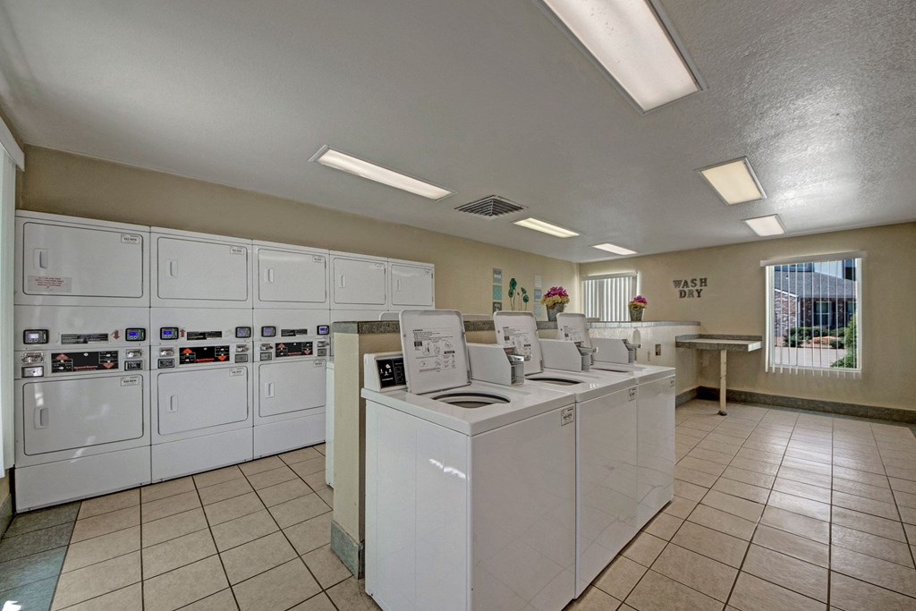 Modern laundromat here at Cypress Pointe Apartment Homes with bright lighting and clean white washers and dryers. Two windows, decorative flowers, and "Wash Dry" on the wall add charm.