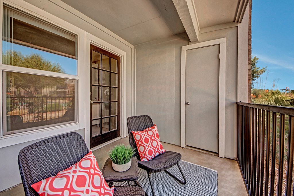 Cozy balcony here at Cypress Pointe Apartment Homes with two wicker chairs adorned with red patterned pillows. A potted plant sits between them on a mat. Glass door and window view greenery under a clear sky.