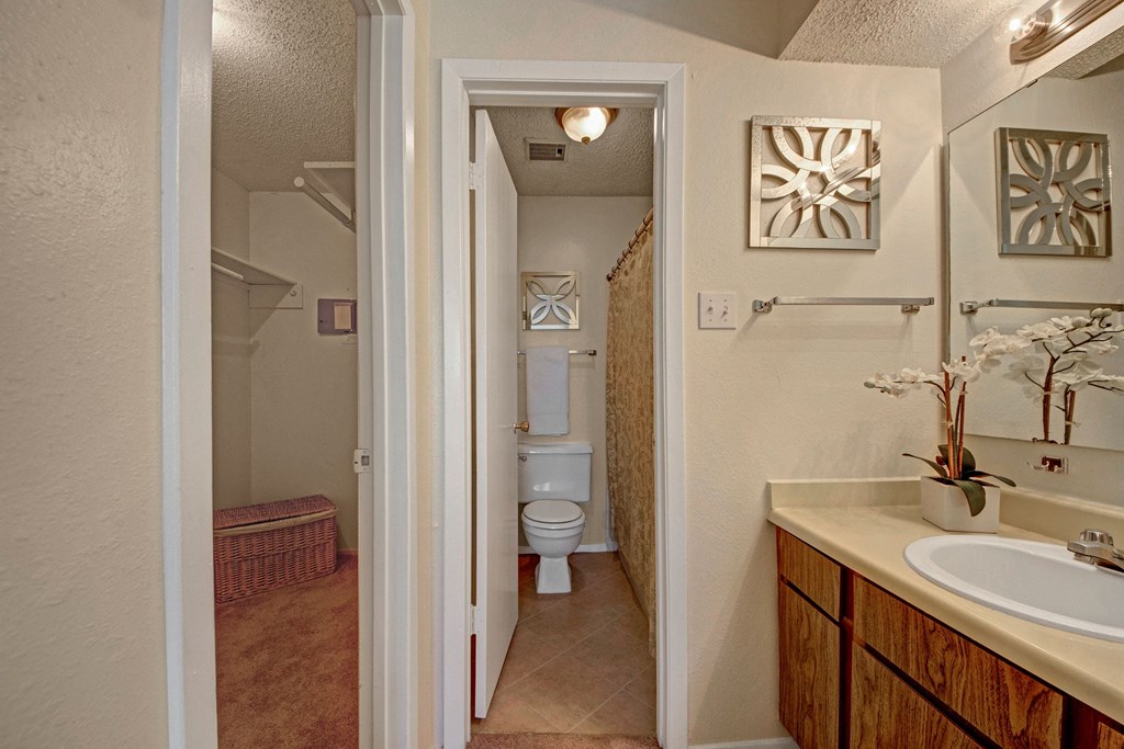 A cozy bathroom and closet scene here at Cypress Pointe Apartment Homes. The bathroom features a toilet, floral shower curtain, ornamental mirror, and an orchid on a vanity. Neutral tones create warmth.