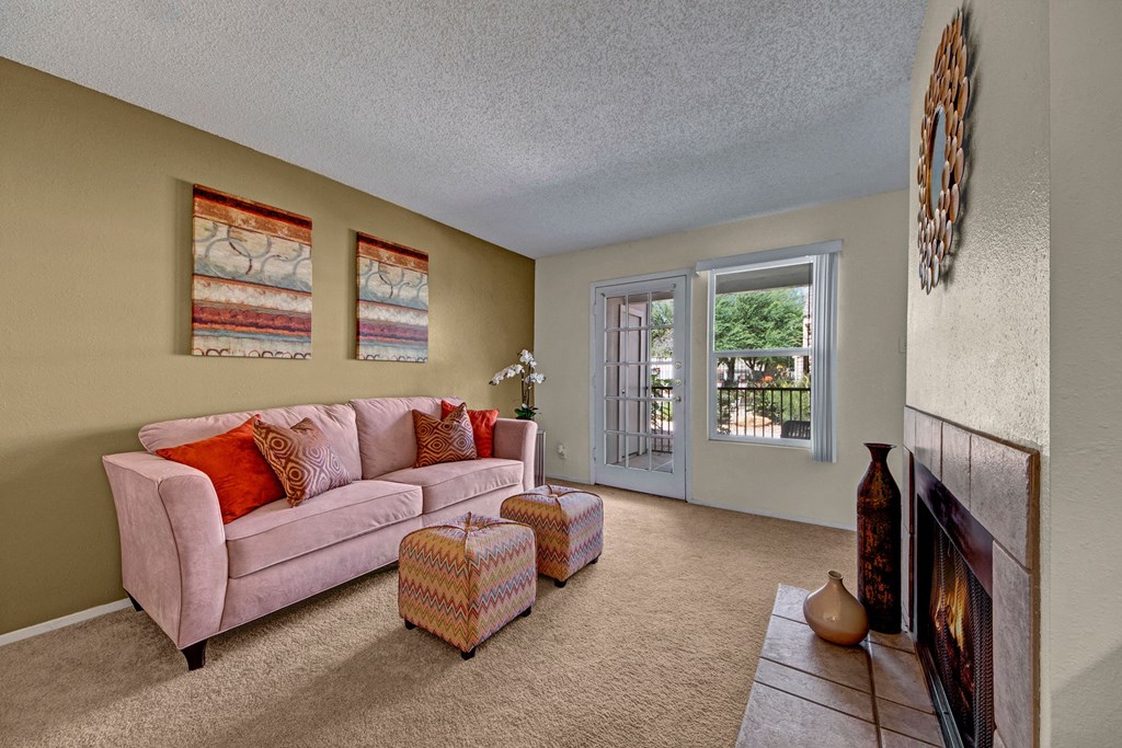 Cozy living room here at Cypress Pointe Apartment Homes with a soft pink sofa, vibrant cushions, and patterned ottomans. Art on the wall, large window, fireplace, and decorative vases add warmth.