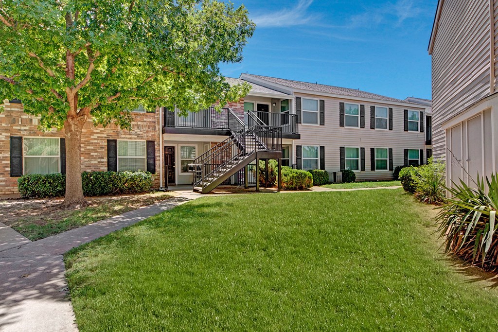Two-story Cypress Pointe Apartment Homes building with tan siding and dark shutters, featuring an outdoor staircase. A lush tree provides shade on a green lawn under a clear blue sky.