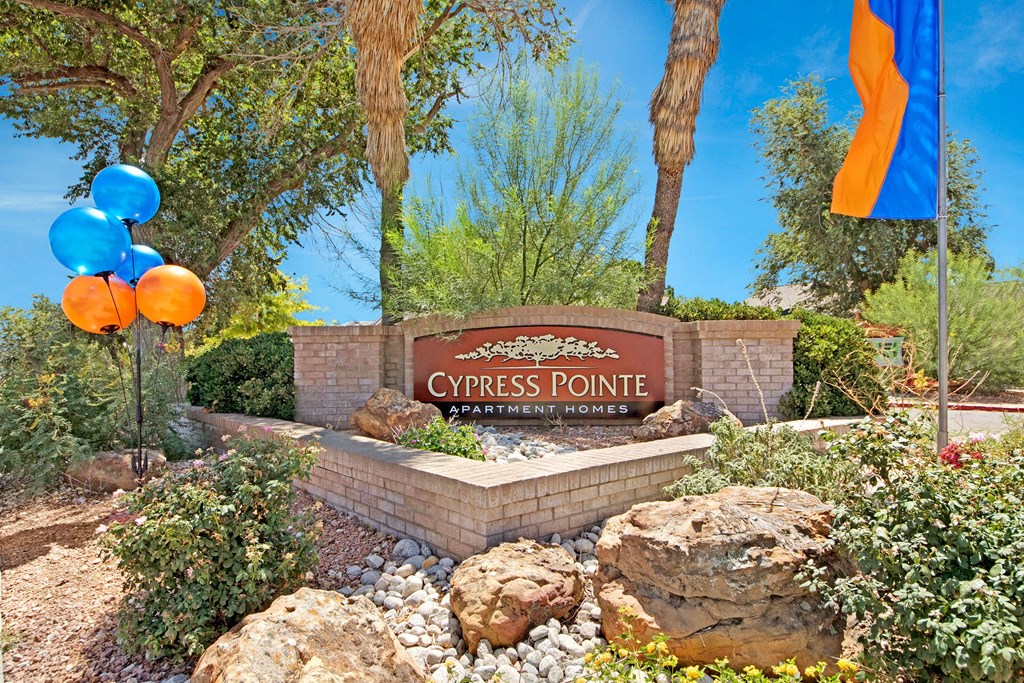 Entrance sign for Cypress Pointe Apartment Homes. Surrounded by lush greenery and rocks, blue and orange balloons float nearby under a sunny sky.