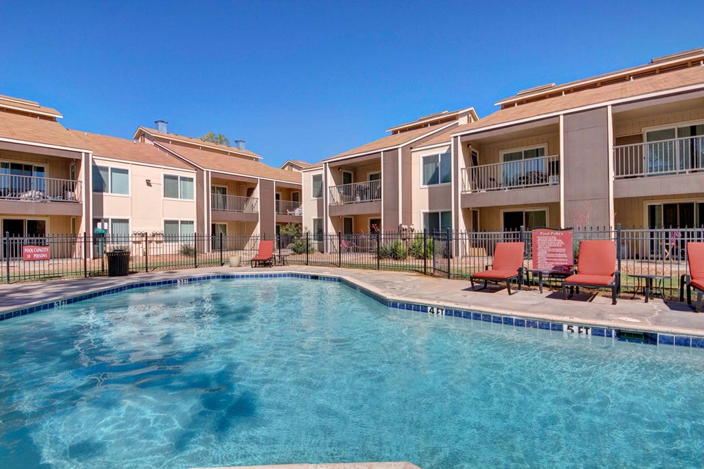 Apartment complex pool area here at Fairgreen Apartment Homes under a clear blue sky, with tan buildings in the background. Red lounge chairs and black fence surround the pool.