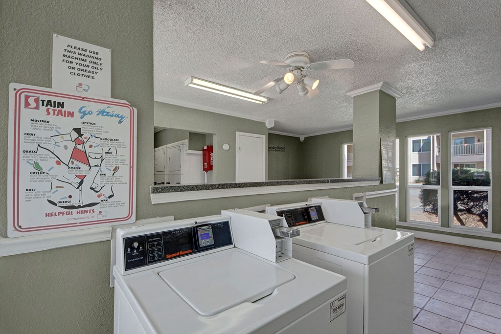 Bright laundry room here at Fairgreen Apartment Homes with two white washing machines, a ceiling fan, and a stain removal guide on the wall. Large windows let in natural light.